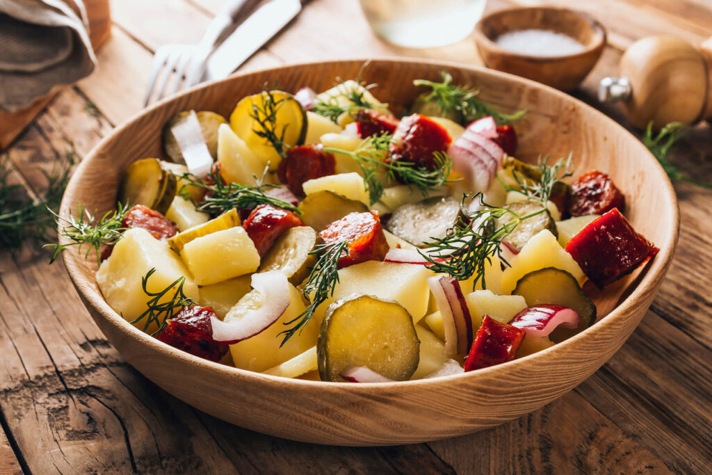 Nahaufnahme des fertigen deutschen Kartoffelsalats ohne Mayonnaise in der Holzschüssel mit glänzenden Kartoffelstücken, knusprig gebratener geräucherter Wurst, Gurkenscheiben, roten Zwiebeln und frischem Dill