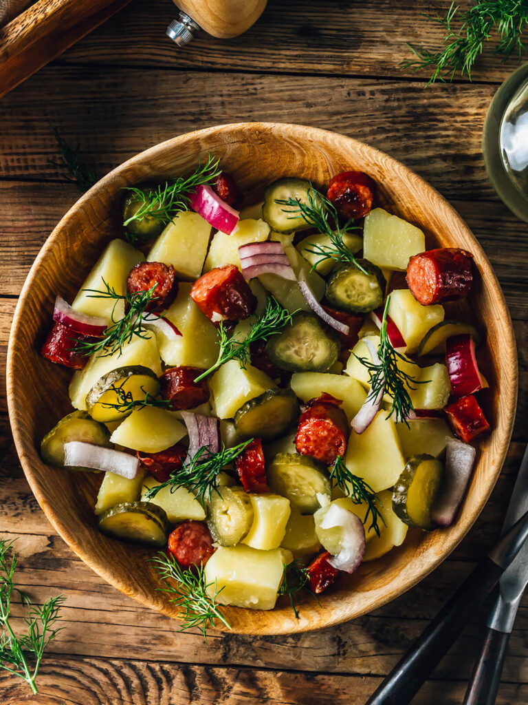 Deutscher Kartoffelsalat ohne Mayonnaise im Hochformat von oben in einer Holzschüssel, mit Essig-Öl-Dressing marinierte Kartoffelstücke, angebratene geräucherte Wurst, Gewürzgurken, rote Zwiebeln und viel frischer Dill