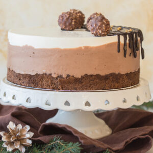 Ganze Schokoladen-Mousse-Torte auf einer weißen Tortenplatte, mit Schokoladenguss und Schoko-Haselnuss-Kugeln dekoriert