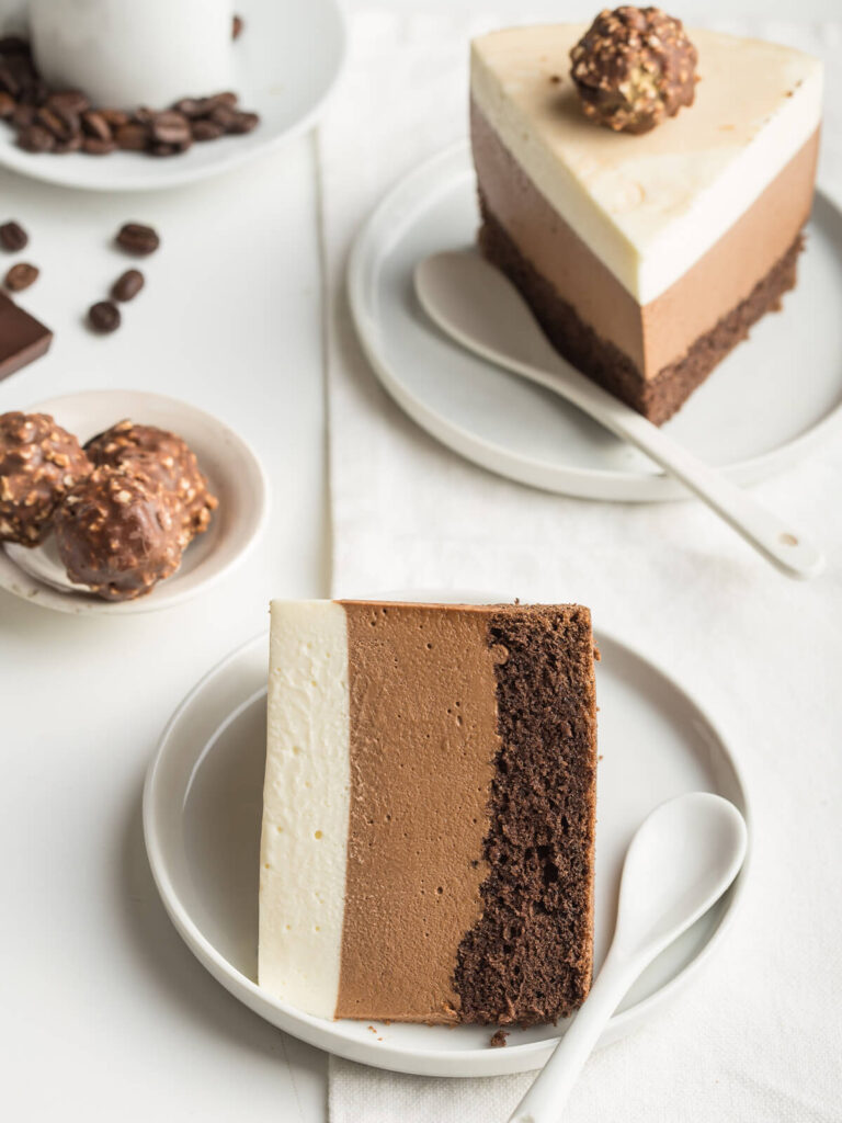 Zwei Stücke Drei-Schokoladen-Mousse-Torte auf Tellern, die drei Mousse-Schichten und der Biskuitboden sind deutlich sichtbar