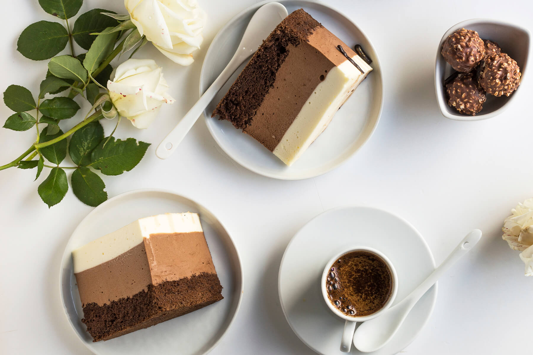 Zwei Stücke Schokoladen-Mousse-Torte von oben, daneben Espresso, weiße Rosen und Schoko-Haselnuss-Kugeln