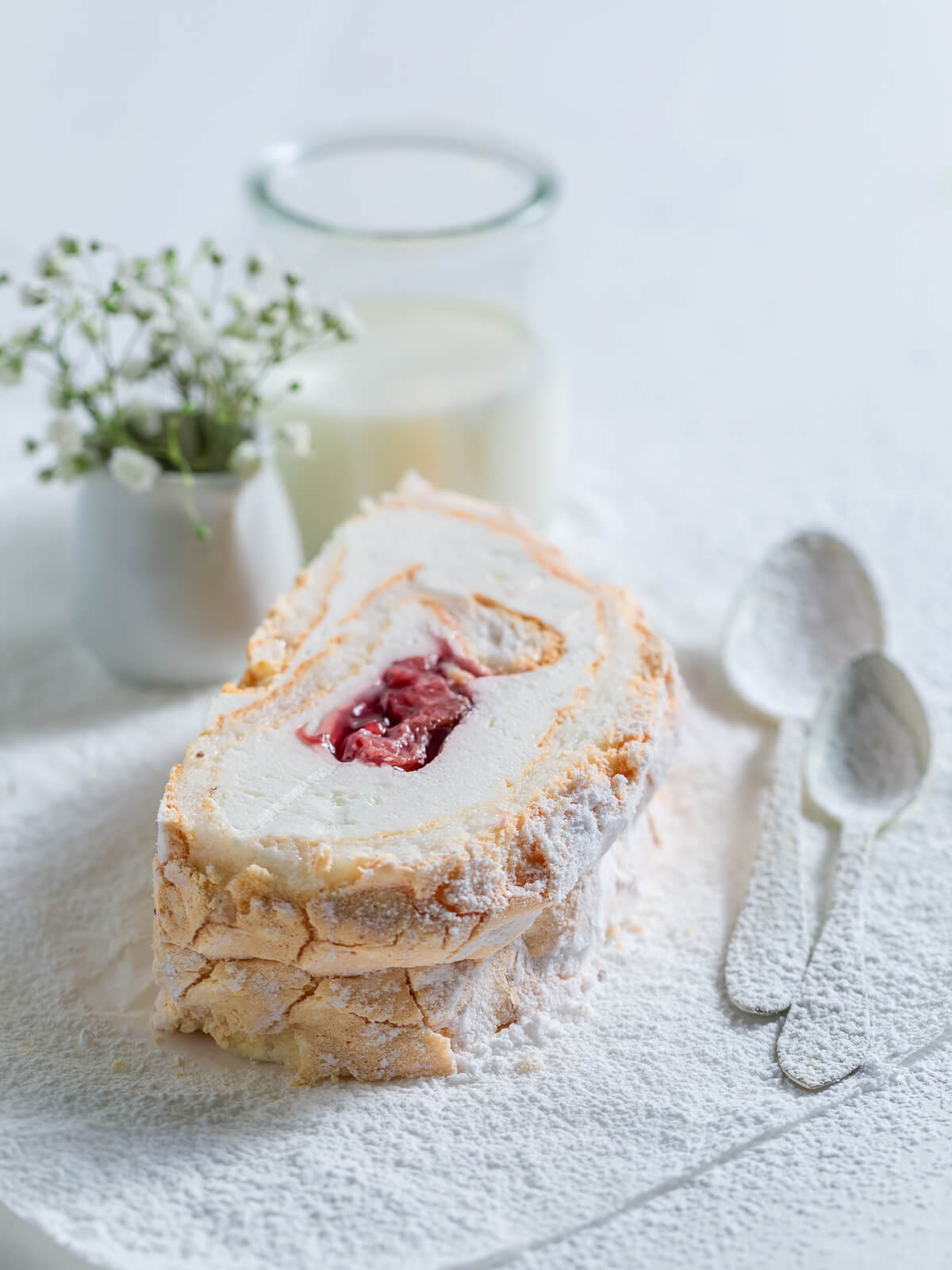 Scheibe Baiser-Roulade mit Sahne und Erdbeer-Füllung neben einem Glas Milch, dick mit Puderzucker bestäubt