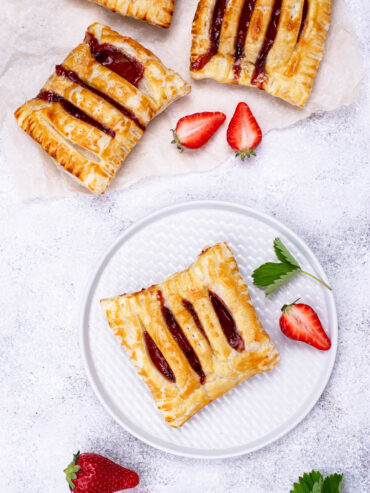 Goldbraune Blätterteig-Taschen mit Erdbeerkonfitüre und eingeschnittener Oberseite, eine auf einem weißen Teller, dazu frische Erdbeeren