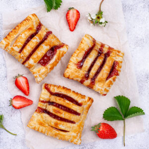 Goldbraune Blätterteig-Taschen mit Erdbeerkonfitüre und eingeschnittener Oberseite auf Backpapier, dazu frische Erdbeeren