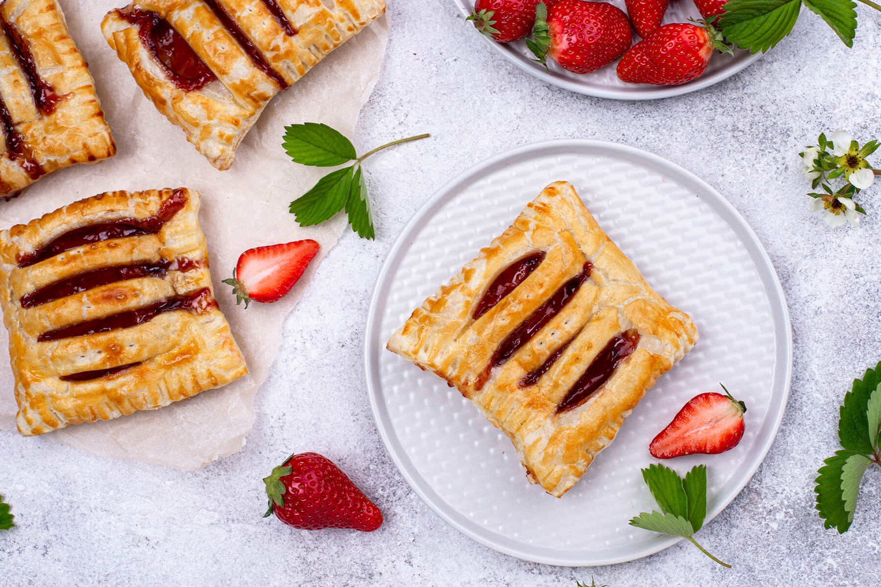 Goldbraune Blätterteig-Taschen mit Erdbeerkonfitüre, teils auf Backpapier und eine auf einem weißen Teller, umgeben von frischen Erdbeeren und Erdbeerblüten