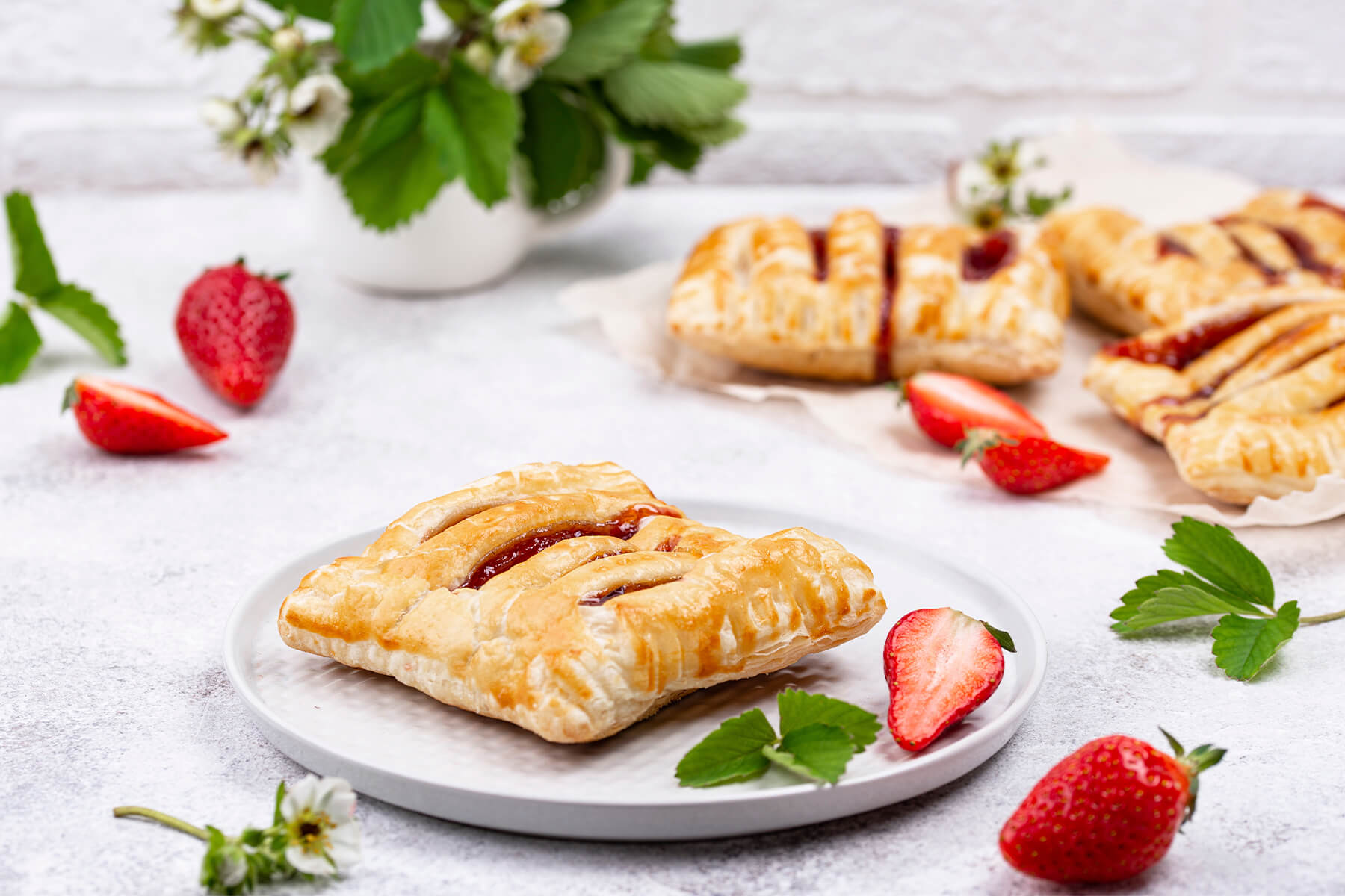 Eine Blätterteig-Tasche mit Erdbeerkonfitüre auf einem weißen Teller, im Hintergrund weitere Taschen und frische Erdbeeren