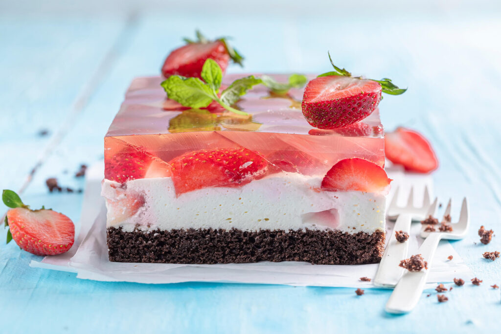 Angeschnittener Blechkuchen mit dunklem Schokoboden, weißer Creme und glasklarem Gelee mit halbierten Erdbeeren auf hellblauem Holz