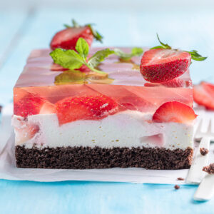 Angeschnittener Blechkuchen mit dunklem Schokoboden, weißer Creme und glasklarem Gelee mit halbierten Erdbeeren auf hellblauem Holz
