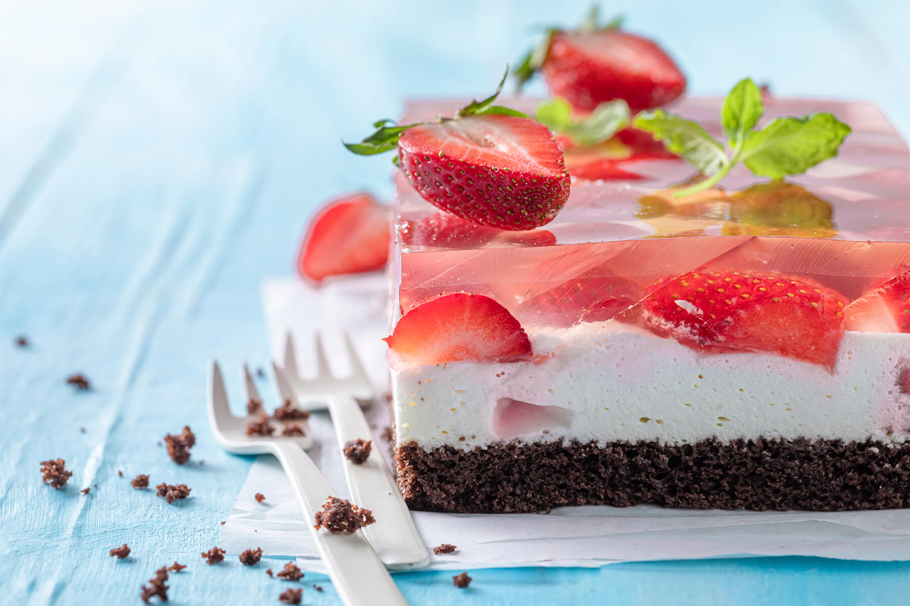 Seitenansicht der Erdbeer-Gelee-Schnitten mit den drei gleich hohen Schichten Schokoboden, weiße Creme und klares Gelee mit eingeschlossenen Erdbeeren
