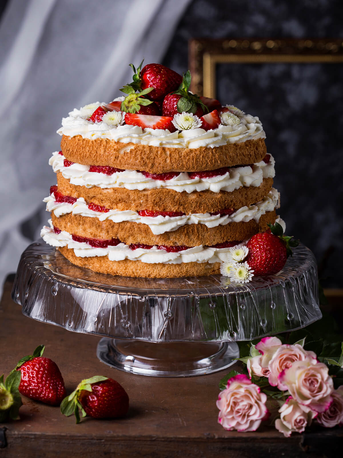 Hohe Erdbeer-Sahne-Torte mit luftigem Biskuit, Sahneschichten und frischen Erdbeeren, garniert mit weißen Blüten, auf einer Glas-Tortenplatte