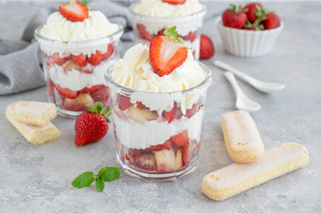 Drei Gläser Erdbeer-Tiramisu mit Sahnehaube und Erdbeeren, daneben Löffelbiskuits und frische Erdbeeren
