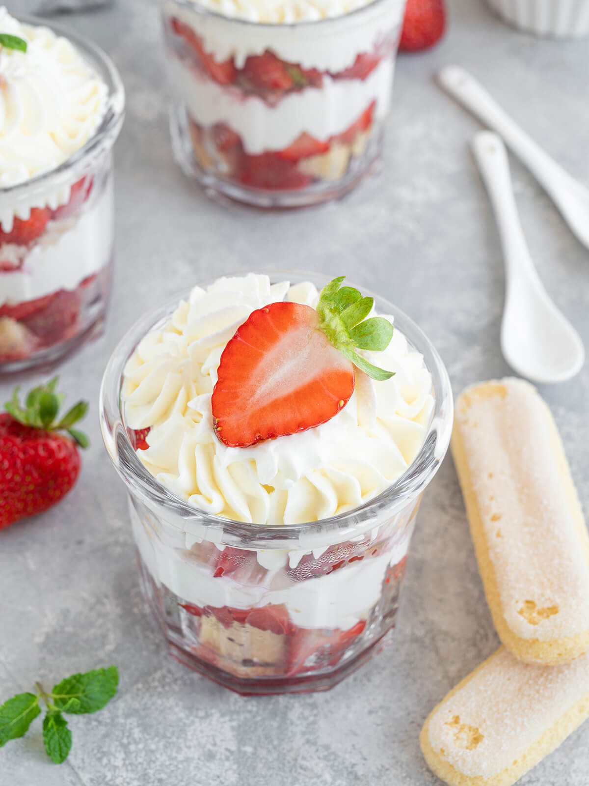 Erdbeer-Tiramisu im Glas von vorn mit sichtbaren Schichten aus Löffelbiskuits, Creme und Erdbeeren, dahinter weitere Gläser und Löffelbiskuits