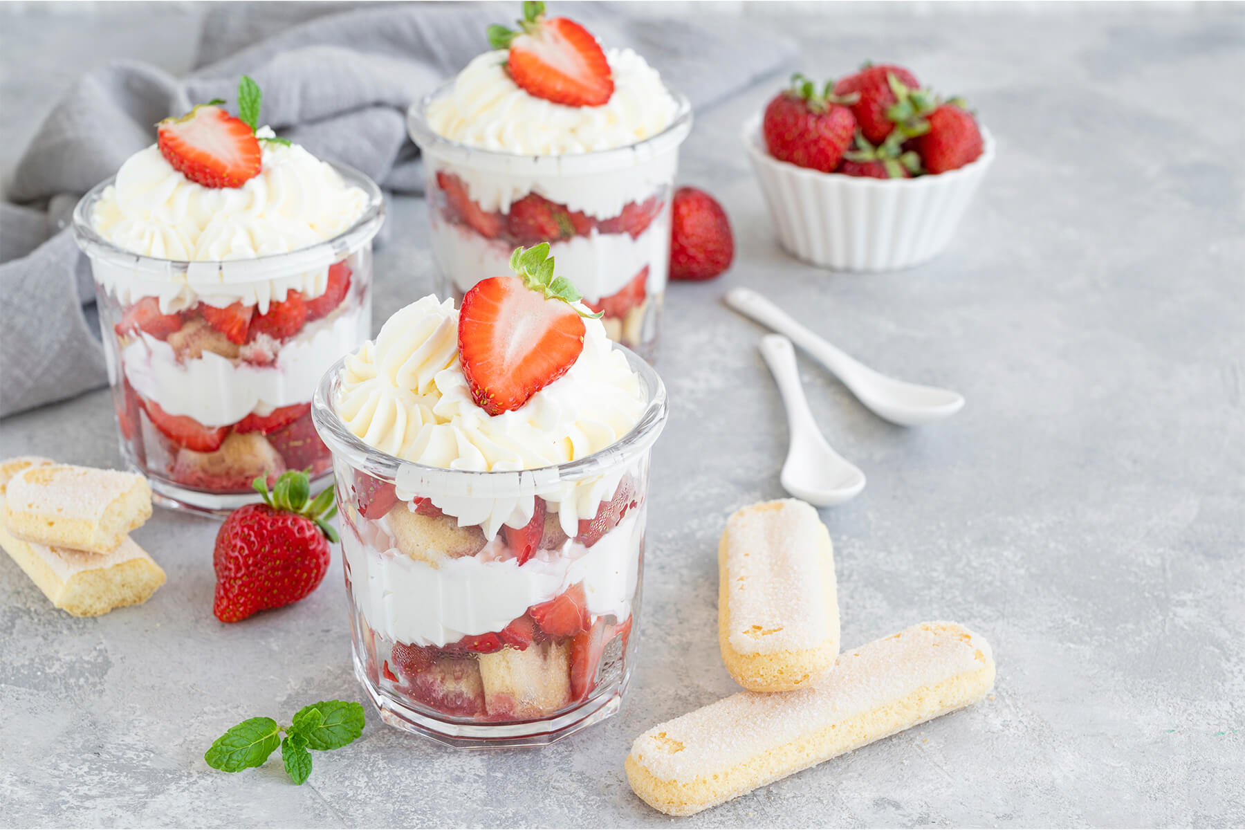 Erdbeer-Tiramisu im Glas mit aufgespritzter Sahnerosette und halbierter Erdbeere, dazu frische Minze