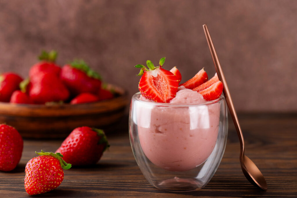 Ein doppelwandiges Glas mit cremiger, zartrosa Erdbeer-Tofu-Mousse, garniert mit halbierten frischen Erdbeeren, daneben ein kupferfarbener Löffel und eine Schale voller Erdbeeren auf einem Holztisch