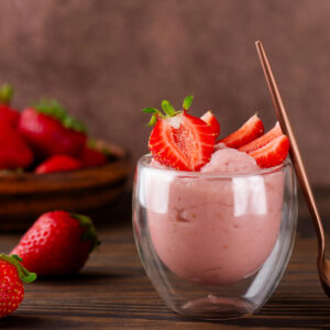 Ein doppelwandiges Glas mit cremiger, zartrosa Erdbeer-Tofu-Mousse, garniert mit halbierten frischen Erdbeeren, daneben ein kupferfarbener Löffel und eine Schale voller Erdbeeren auf einem Holztisch