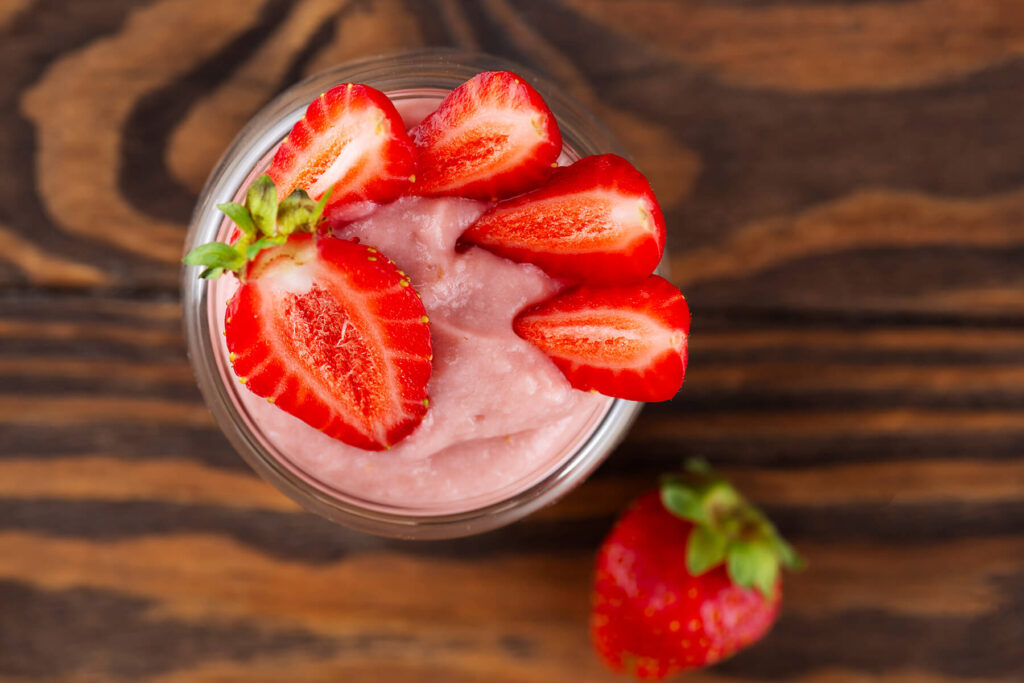 Aufsicht auf ein Glas cremige, zartrosa Erdbeer-Tofu-Mousse, rundum garniert mit halbierten frischen Erdbeeren, auf dunklem Holz