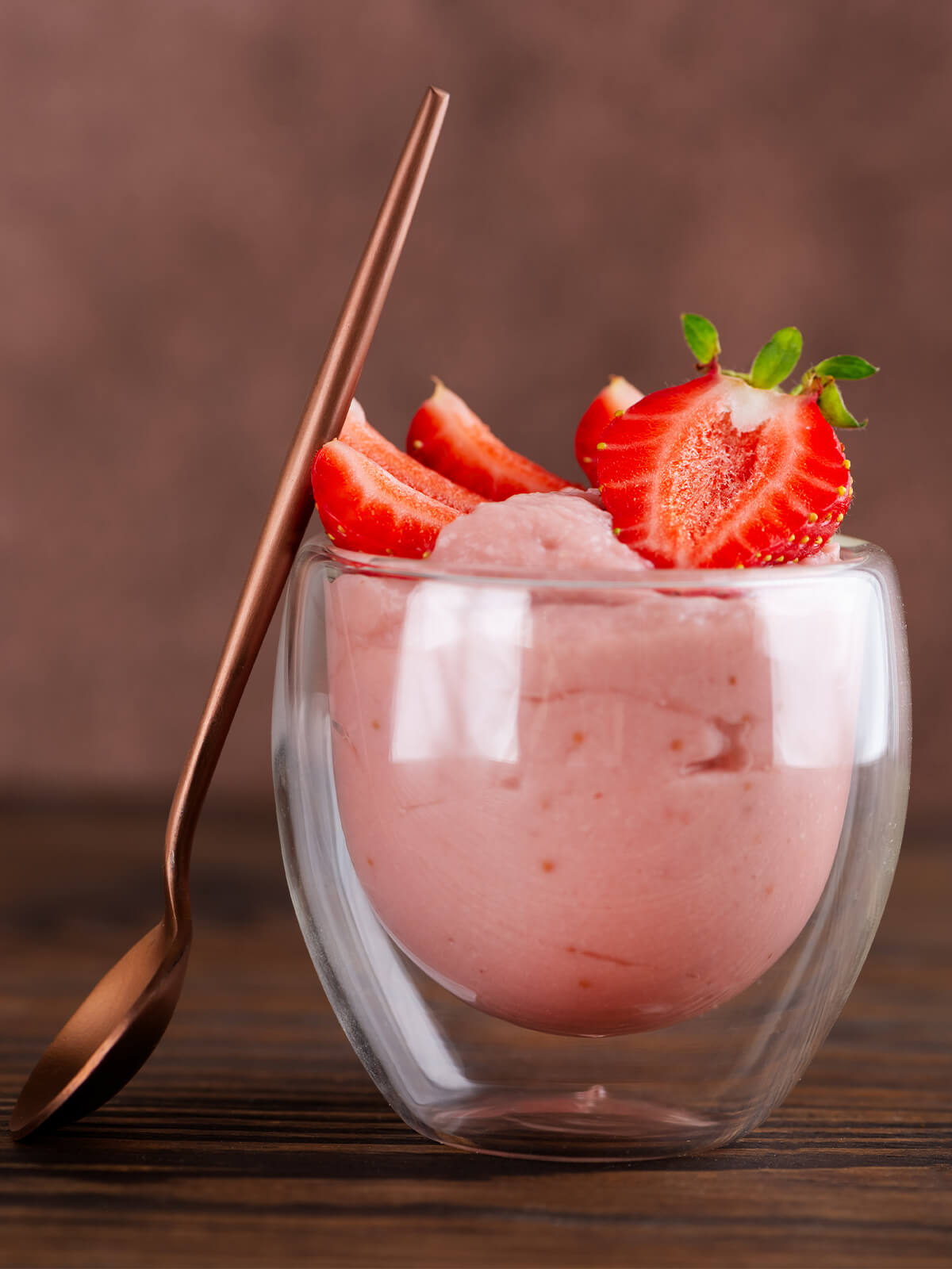 Ein doppelwandiges Glas mit seidig-glatter, zartrosa Erdbeer-Tofu-Mousse, obenauf halbierte und geviertelte frische Erdbeeren, daneben ein kupferfarbener Löffel vor warmem braunem Hintergrund
