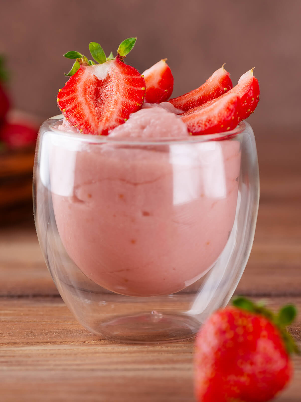Ein doppelwandiges Glas mit cremiger, zartrosa Erdbeer-Tofu-Mousse und frischen halbierten Erdbeeren obenauf, im Hintergrund eine Schale mit Erdbeeren auf Holz