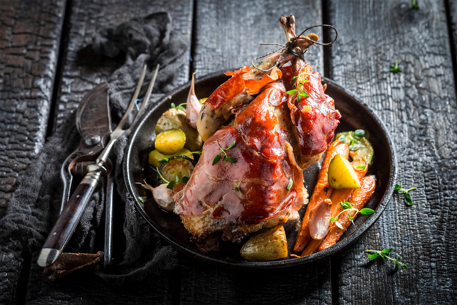 Gebratener Fasan im Rohschinkenmantel mit Karotten, Kartoffeln und Knoblauch in einer Pfanne, daneben Tranchierbesteck