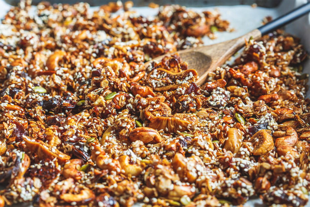 Kerniges Feigen-Dattel-Granola mit Cashews, Kürbiskernen und Sesam auf einem Backblech, dazu ein Holzlöffel