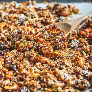 Kerniges Feigen-Dattel-Granola mit Cashews, Kürbiskernen und Sesam auf einem Backblech, dazu ein Holzlöffel