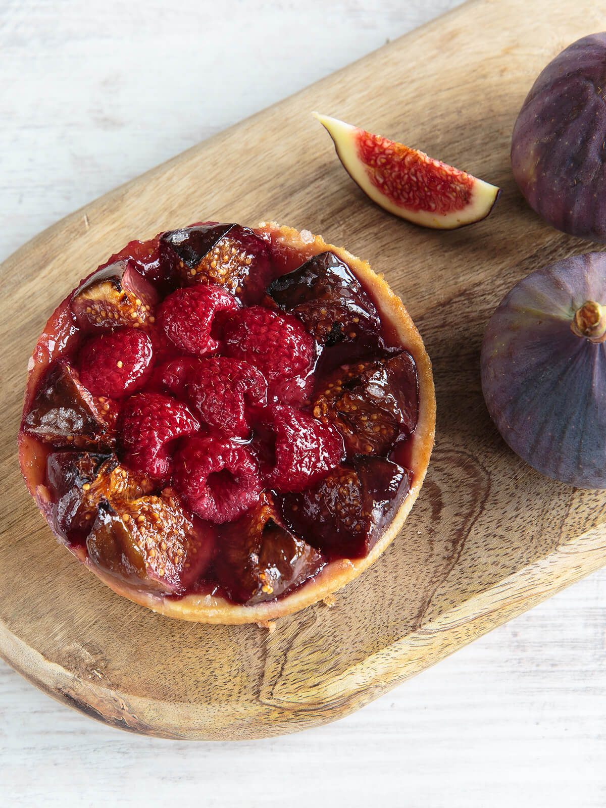 Feigen-Himbeer-Tartelette mit dunkel gebackenen Feigenspalten und mitgebackenen Himbeeren, daneben frische Feigen auf hellem Holz