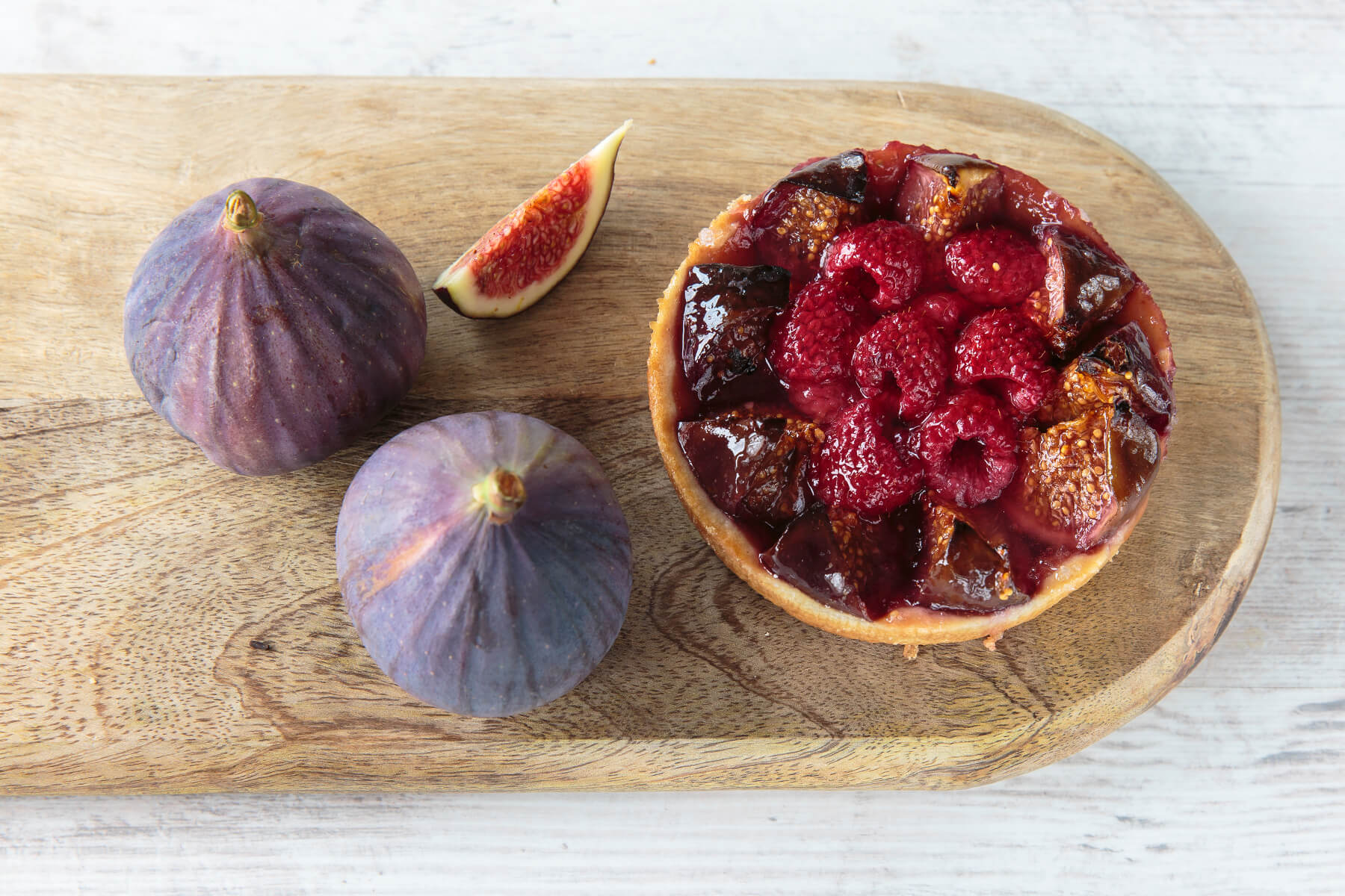 Feigen-Himbeer-Tartelette mit gebackenen Feigenspalten und dunklen Himbeeren, daneben zwei ganze Feigen und ein Feigenstück