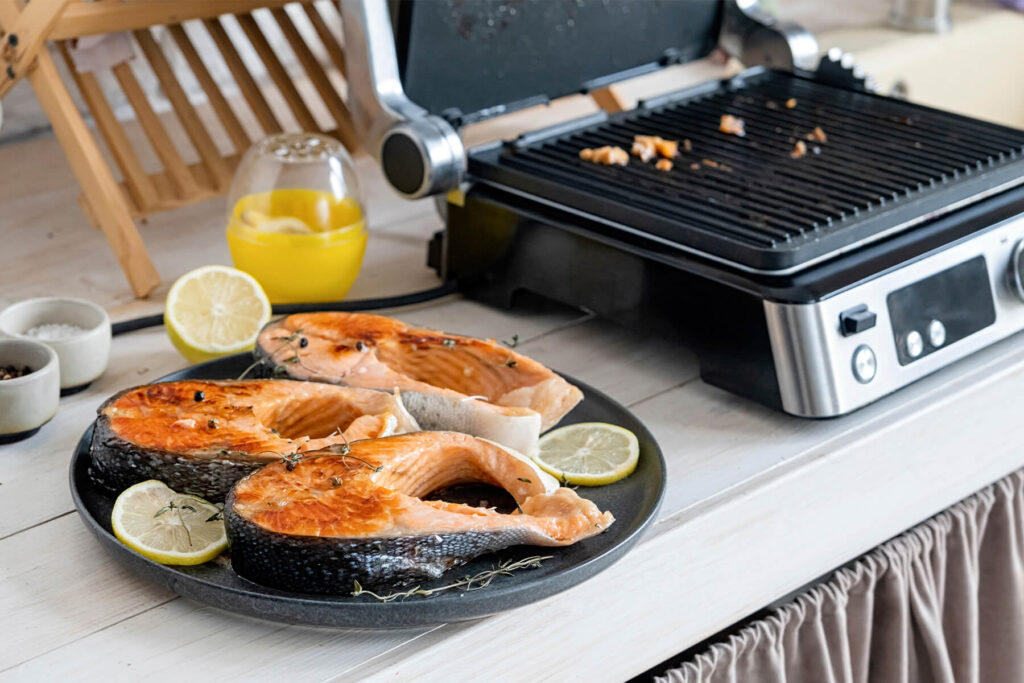 Gegrillte Forellensteaks mit goldbrauner Kruste, Zitronenscheiben und Thymian auf einem dunklen Teller neben dem Kontaktgrill
