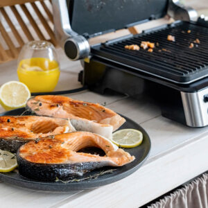 Gegrillte Forellensteaks mit goldbrauner Kruste, Zitronenscheiben und Thymian auf einem dunklen Teller neben dem Kontaktgrill