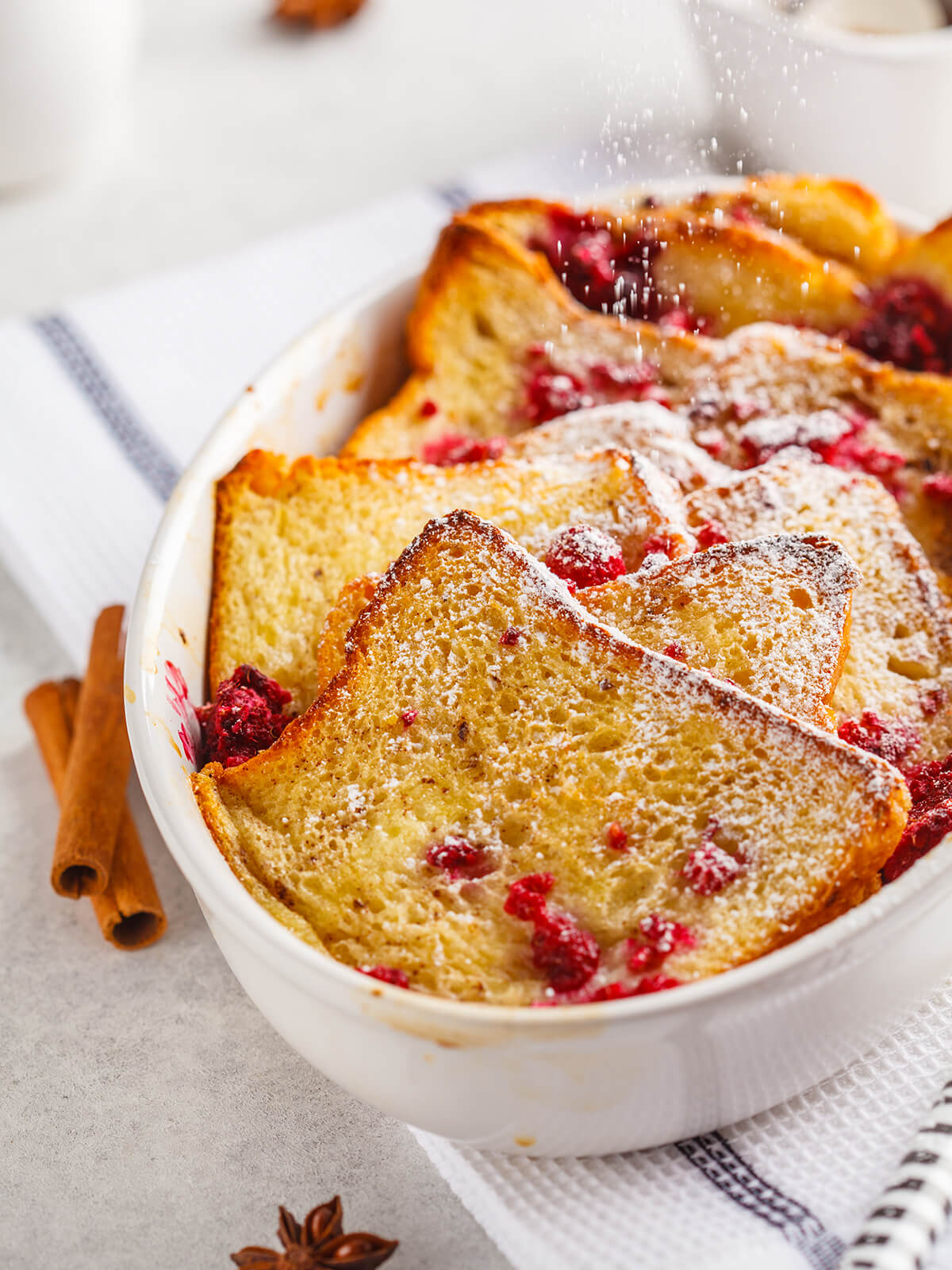 Goldbrauner French-Toast-Auflauf mit Himbeeren, der mit Puderzucker bestäubt wird, in einer weißen Auflaufform mit Zimtstangen und Sternanis