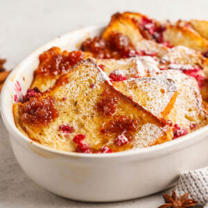 Goldbrauner French-Toast-Auflauf mit Himbeeren, Himbeermarmelade und Puderzucker in einer weißen Auflaufform, daneben Zimtstangen