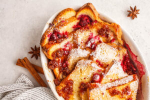 French-Toast-Auflauf von oben in einer weißen Form mit Himbeeren, Himbeermarmelade und Puderzucker, daneben Zimtstangen und Sternanis