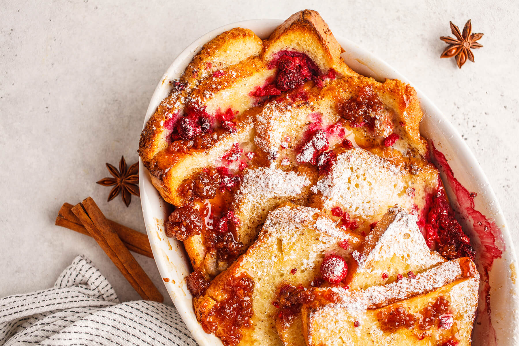 French-Toast-Auflauf von oben in einer weißen Form mit Himbeeren, Himbeermarmelade und Puderzucker, daneben Zimtstangen und Sternanis