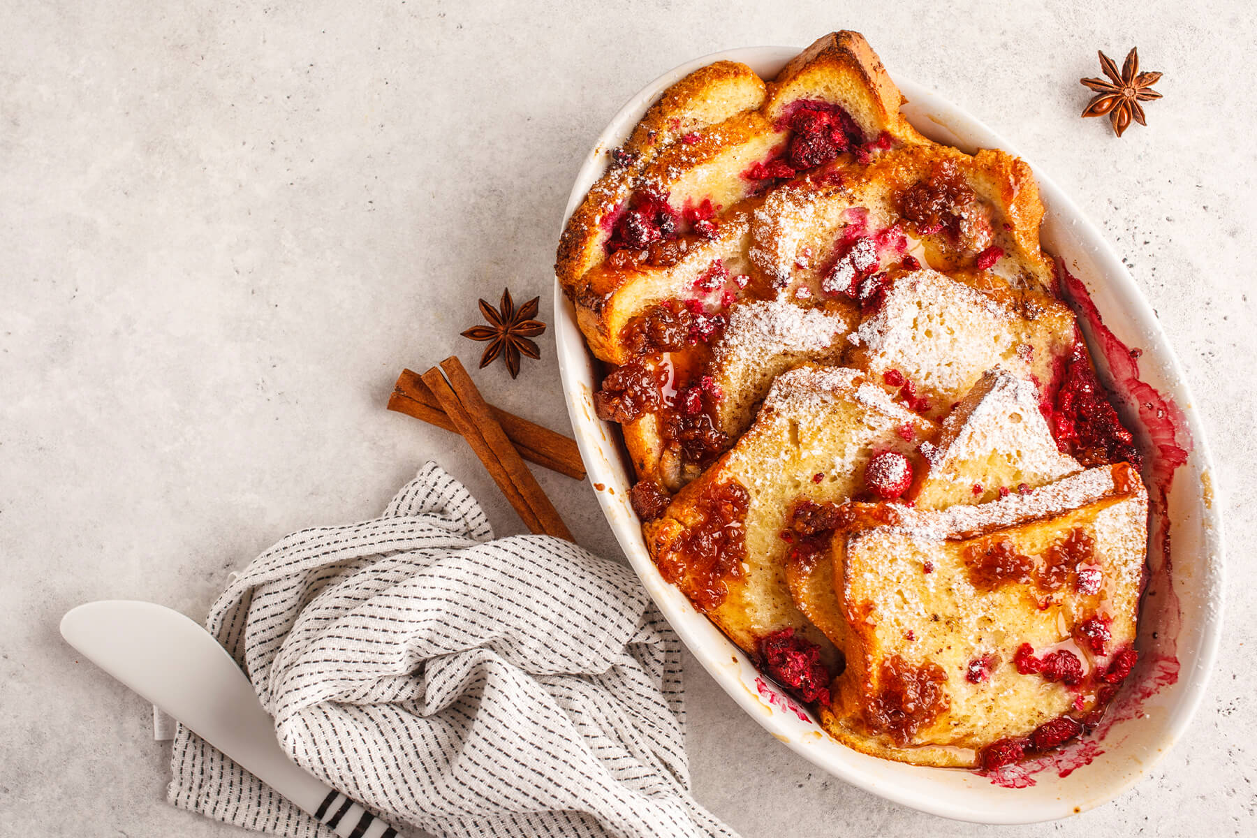 French-Toast-Auflauf mit Himbeeren und Puderzucker in einer weißen Auflaufform von oben, daneben ein Servierheber und ein gestreiftes Tuch