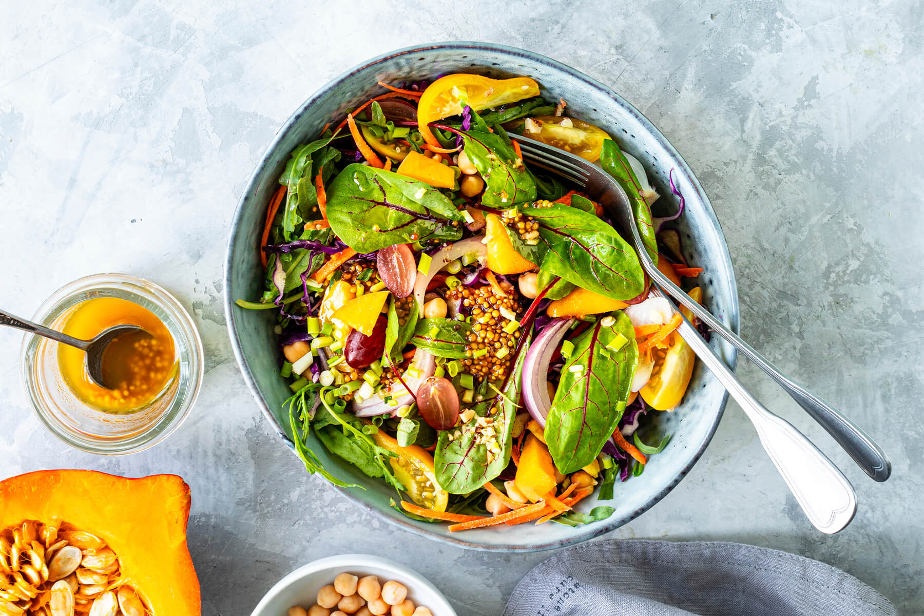 Bunter Blattsalat mit rohem Kürbis, Rotkohl, Kichererbsen, roten Trauben und Senfkörnern von oben, daneben ein Glas Dressing und ein halber Hokkaido-Kürbis