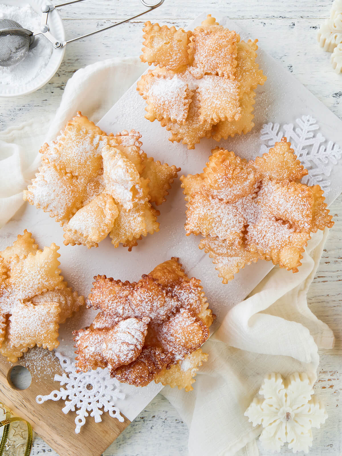 Fünf frittierte Schneeflocken aus gezackten Teigquadraten mit Puderzucker auf einem Marmorbrett, daneben weihnachtliche Deko