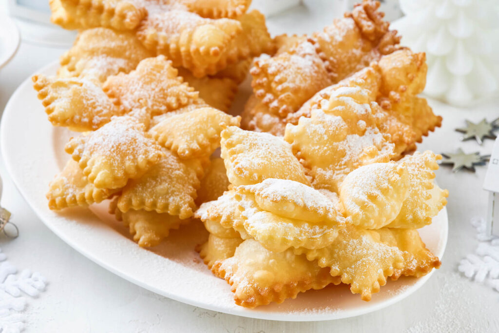 Fertige frittierte Schneeflocken mit aufgestellten, blasig gewölbten Teigblättern und Puderzucker auf einem weißen Teller