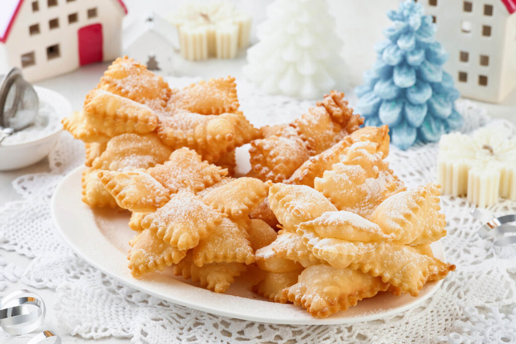 Frittierte Schneeflocken aus gezackten Teigquadraten mit Puderzucker auf einem weißen Teller vor weihnachtlicher Deko
