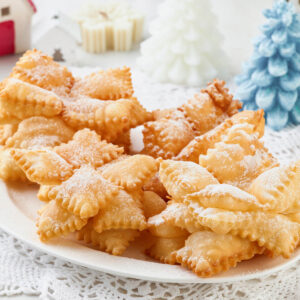Frittierte Schneeflocken aus gezackten Teigquadraten mit Puderzucker auf einem weißen Teller vor weihnachtlicher Deko