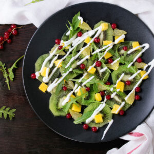 Fruchtsalat mit Rucola von oben auf einem matten schwarzen Teller: dünne Kiwischeiben liegen überlappend als Unterlage und stecken zugleich oben zwischen den Blättern, dazwischen goldgelbe Mangowürfel, blassgelbe durchscheinende Grapefruitstücke, schmale Blätter von wildem Rucola und einzeln verteilte rote Johannisbeeren; darüber läuft eine Zickzacklinie aus dicken weißen Joghurtraupen, die auch auf dem blanken Teller ihre Form behalten; auf dem dunklen Holztisch links Rucolablätter und Rispen roter Johannisbeeren, rechts ein weißes Leinentuch