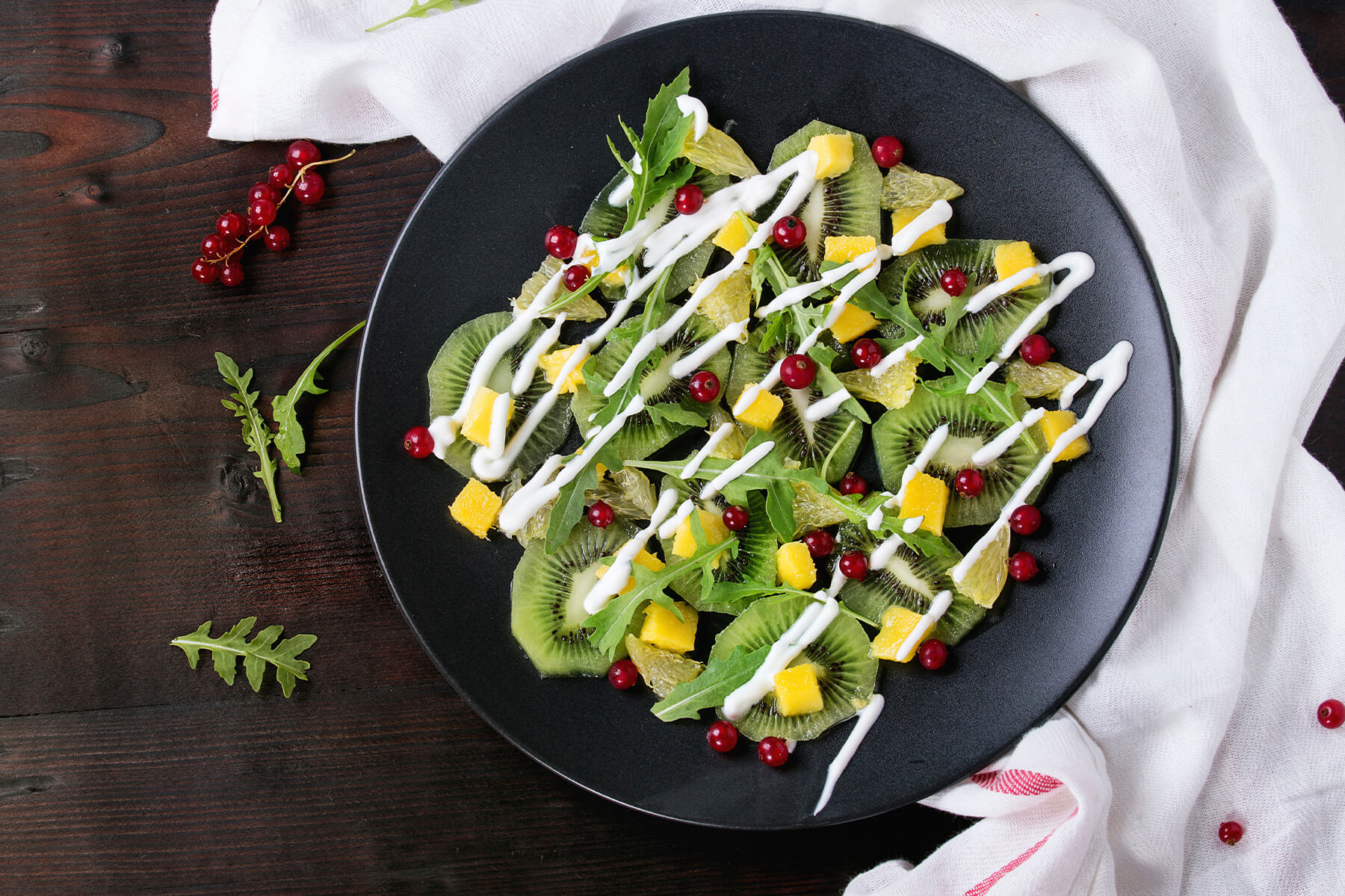 Fruchtsalat mit Rucola von oben auf einem matten schwarzen Teller: dünne Kiwischeiben liegen überlappend als Unterlage und stecken zugleich oben zwischen den Blättern, dazwischen goldgelbe Mangowürfel, blassgelbe durchscheinende Grapefruitstücke, schmale Blätter von wildem Rucola und einzeln verteilte rote Johannisbeeren; darüber läuft eine Zickzacklinie aus dicken weißen Joghurtraupen, die auch auf dem blanken Teller ihre Form behalten; auf dem dunklen Holztisch links Rucolablätter und Rispen roter Johannisbeeren, rechts ein weißes Leinentuch