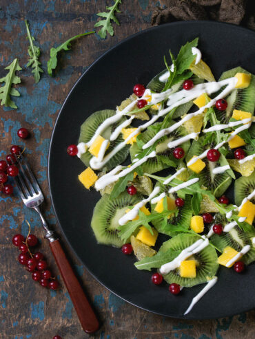 Nahaufnahme eines Fruchtsalats mit Rucola auf schwarzem Teller: überlappende Kiwischeiben mit hellem Kern und Kranz aus schwarzen Kernen, teils als Unterlage, teils oben zwischen den Blättern, dazu goldgelbe Mangowürfel, blassgelbe durchscheinende Grapefruitstücke, schmale Blätter von wildem Rucola und einzelne rote Johannisbeeren; quer darüber laufen etwa fünf Millimeter dicke weiße Joghurtraupen, die bis auf den blanken Teller reichen und dort als Linie stehen bleiben; links auf verwittertem, blau abblätterndem Holz eine Gabel mit dunklem Holzgriff und Rispen roter Johannisbeeren