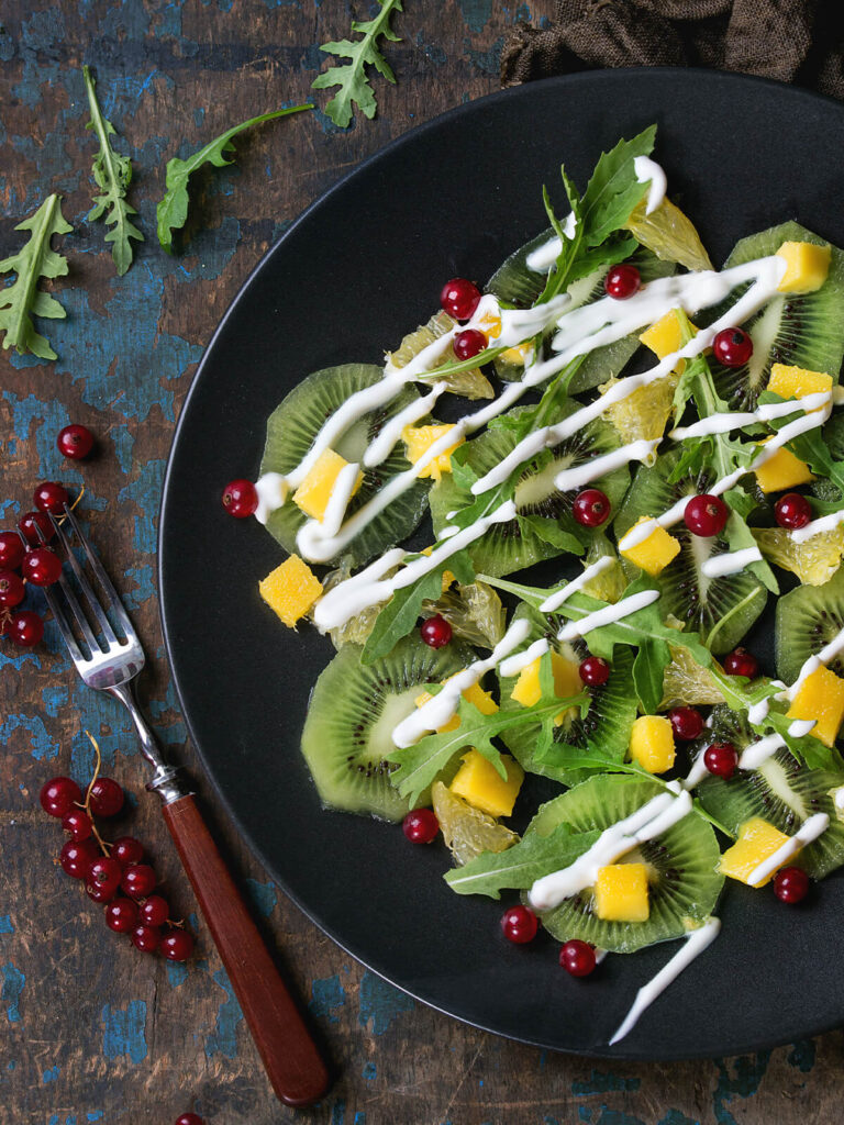 Nahaufnahme eines Fruchtsalats mit Rucola auf schwarzem Teller: überlappende Kiwischeiben mit hellem Kern und Kranz aus schwarzen Kernen, teils als Unterlage, teils oben zwischen den Blättern, dazu goldgelbe Mangowürfel, blassgelbe durchscheinende Grapefruitstücke, schmale Blätter von wildem Rucola und einzelne rote Johannisbeeren; quer darüber laufen etwa fünf Millimeter dicke weiße Joghurtraupen, die bis auf den blanken Teller reichen und dort als Linie stehen bleiben; links auf verwittertem, blau abblätterndem Holz eine Gabel mit dunklem Holzgriff und Rispen roter Johannisbeeren