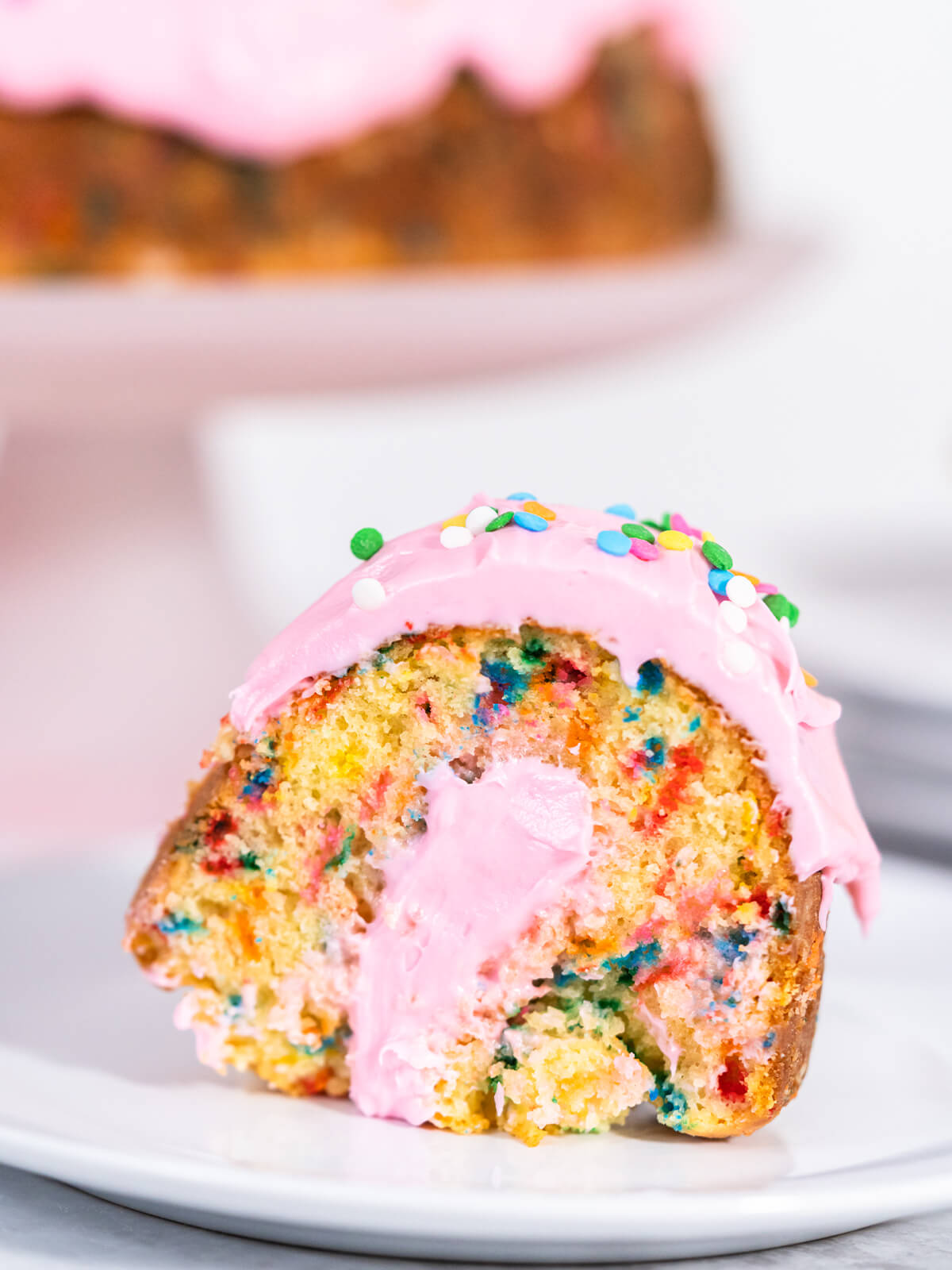 Ein Stück Konfetti-Gugelhupf mit rosa Buttercreme auf einem weißen Teller, im Hintergrund der ganze Kuchen