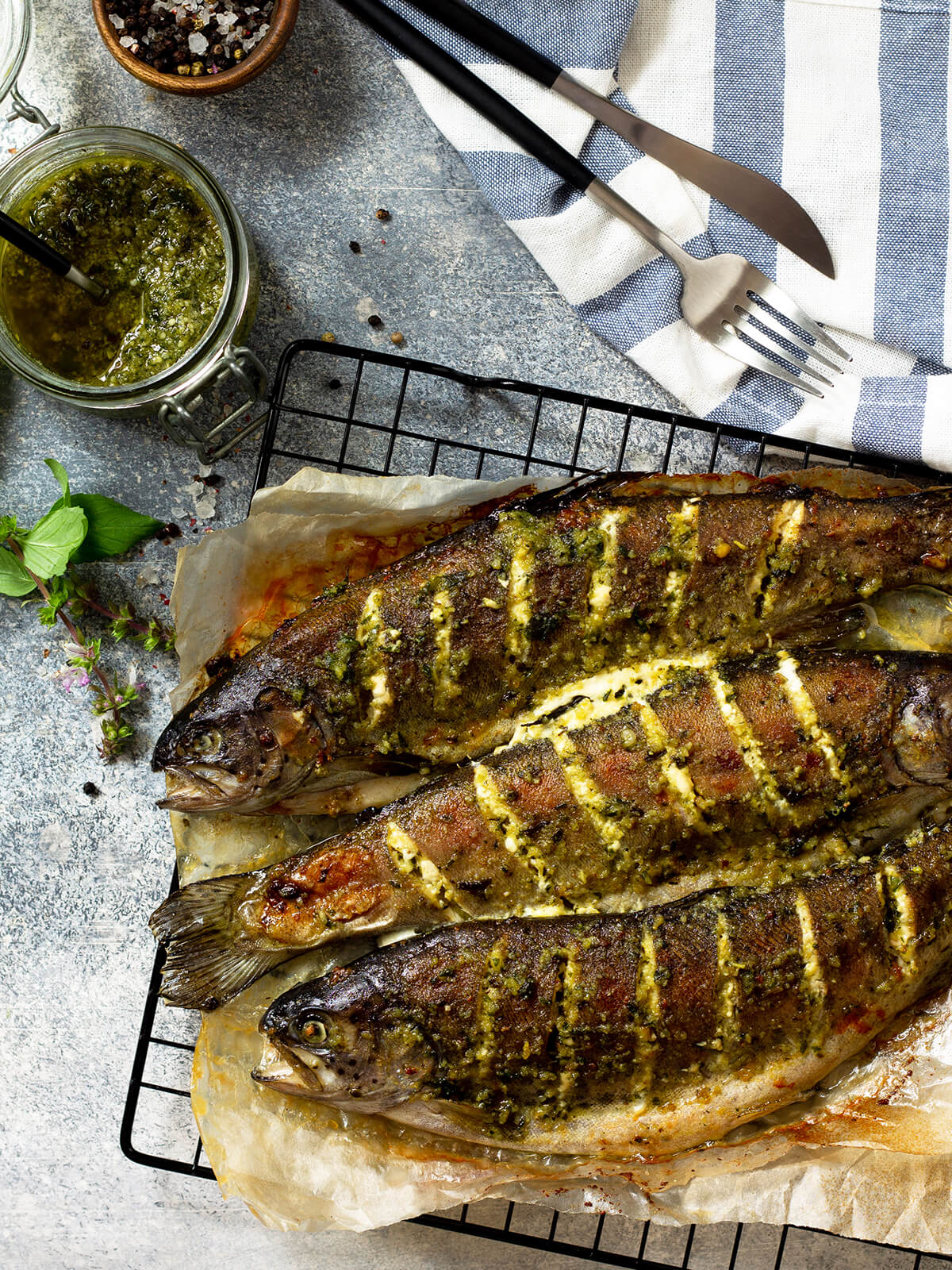 "Drei ganze Forellen mit Pesto von oben auf einem Gitterrost, daneben ein Glas grünes Pesto und frische Kräuter"