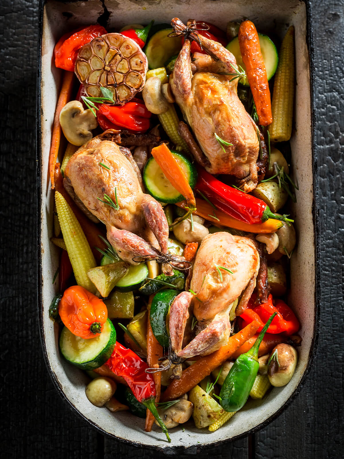 Drei gebackene Wachteln mit gebundenen Keulen auf buntem Ofengemüse aus Karotten, Zucchini, Paprika und Maiskölbchen in einer gusseisernen Form von oben