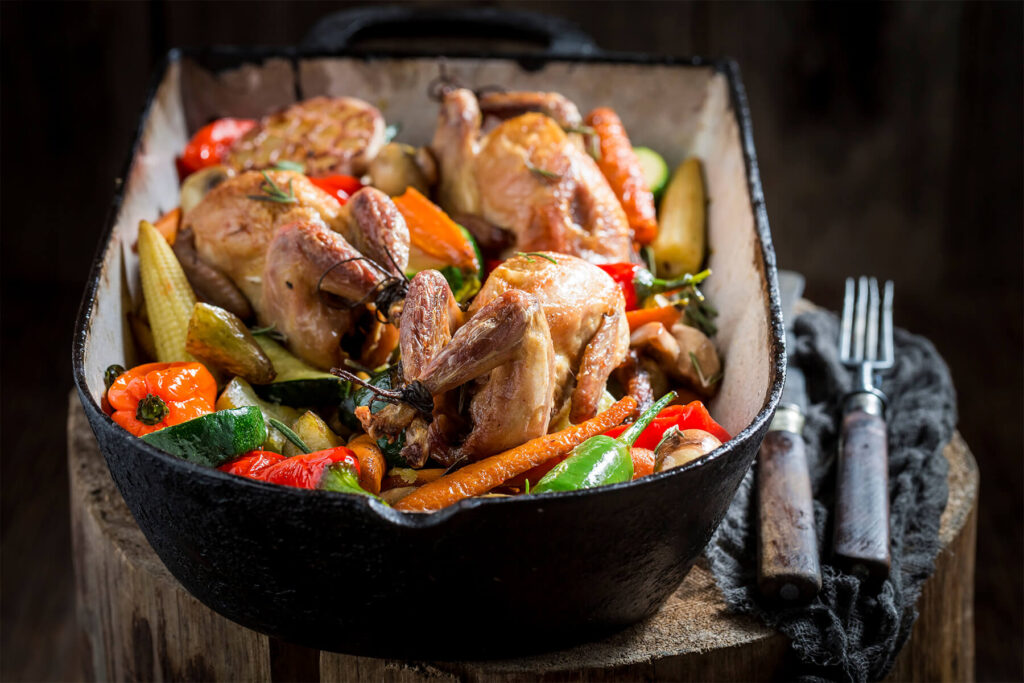 Gebackene Wachteln mit buntem Ofengemüse aus Karotten, Zucchini, Paprika, Maiskölbchen und Champignons in einer gusseisernen Form
