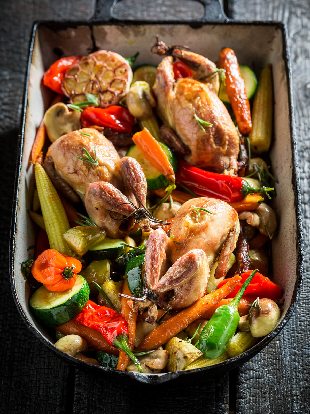 Gebackene Wachteln mit Zucchini, ganzen Karotten, Mini-Paprika, weißen Champignons und geröstetem Knoblauch in einer dunklen Auflaufform