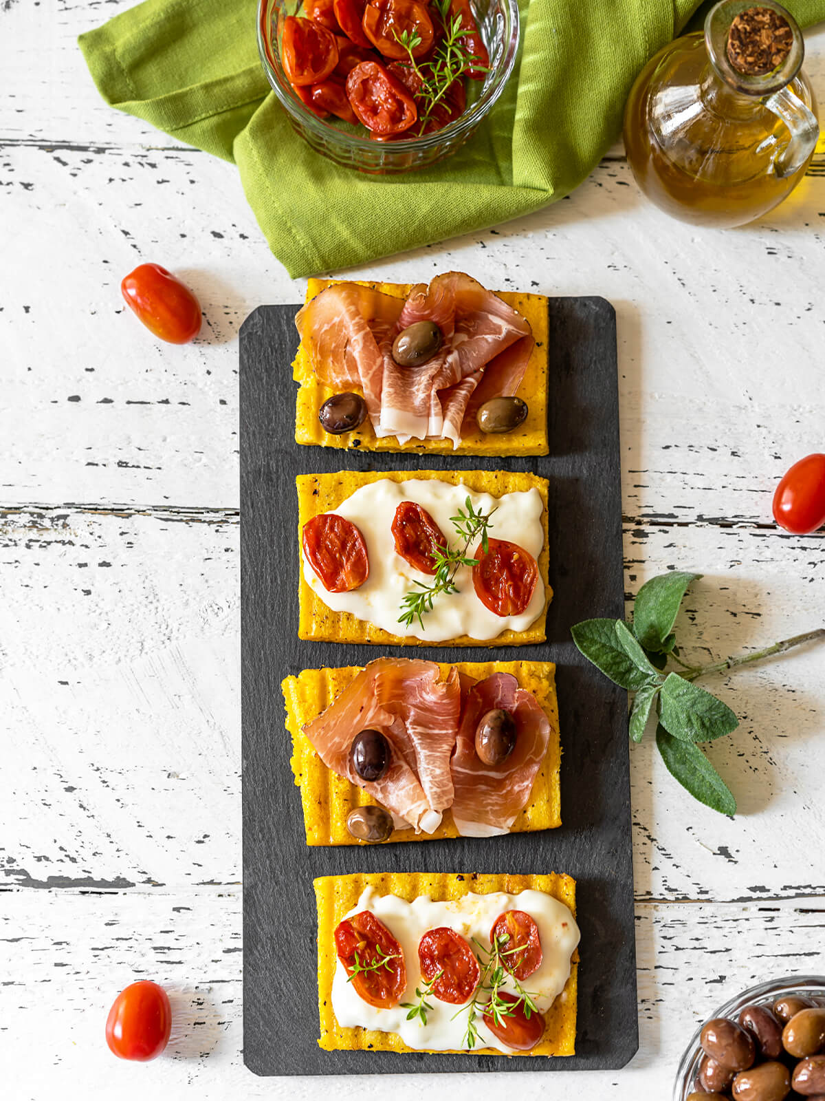 Gegrillte Polenta-Schnitten auf einer Schieferplatte, belegt mit Stracchino und Tomaten sowie mit Prosciutto und Oliven