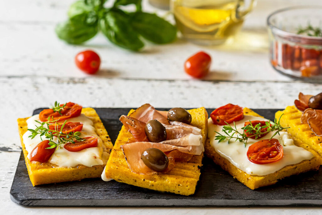 Gegrillte Polenta-Schnitten mit Stracchino, geschmorten Kirschtomaten, Prosciutto und Oliven auf einer Platte